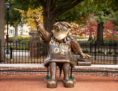 Cocky Statue sitting on a bench with his hand raised doing a spurs up gesture and fall trees in the background. 