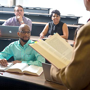 Law students sitting in a classroom watching a professor reading from a law book. 