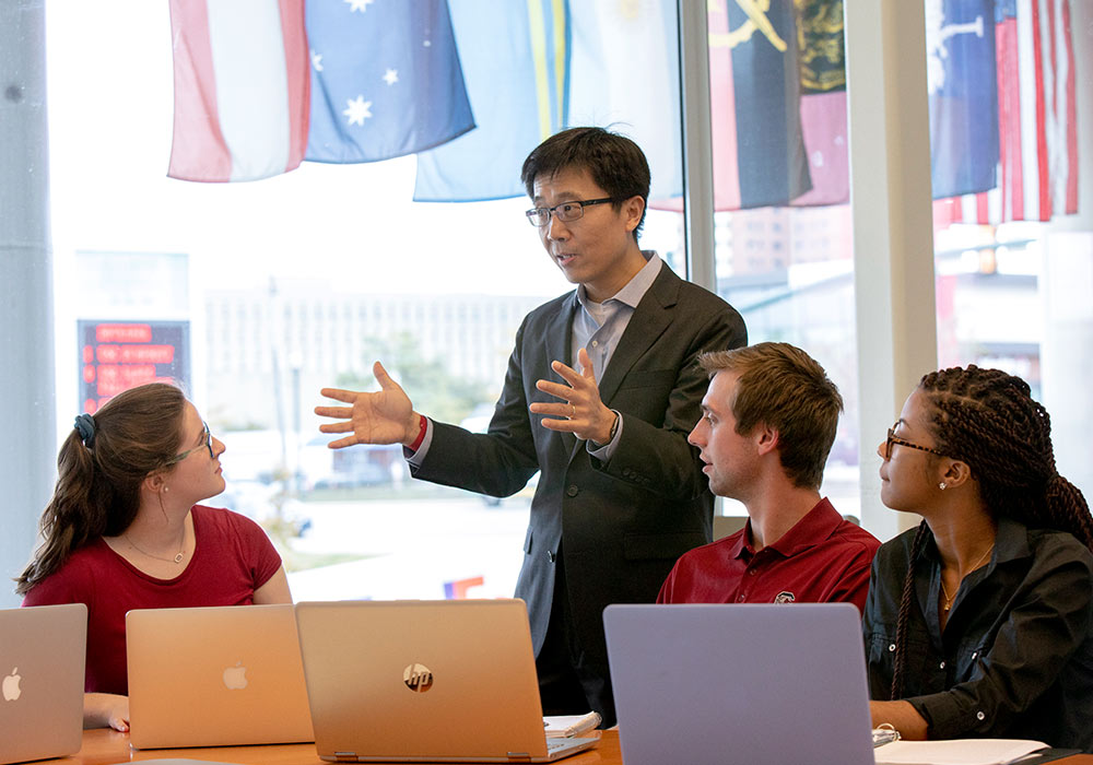 Professor talks with a table of students in the Moore School building with international flags displayed outside the window. 