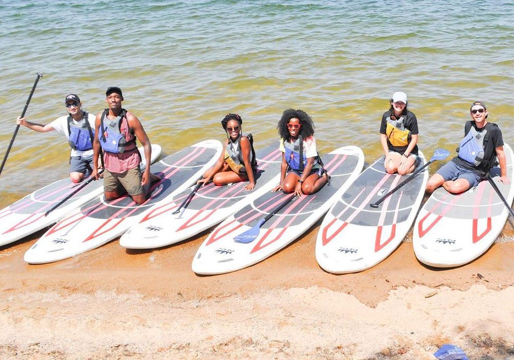 Several people kneeling on paddle boards lined up on the shore of Lake Murray. 