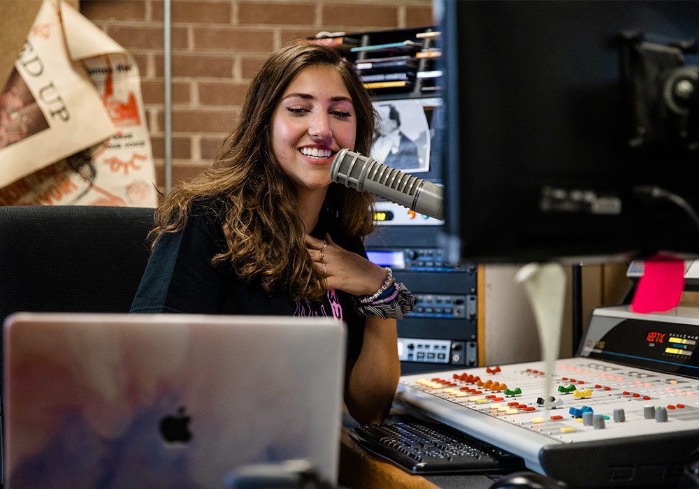 Student DJ behind radio console speaking into a microphone. 