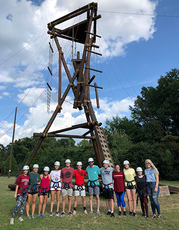 A University 101 class gathered together wearing helmets in front of the rope course
