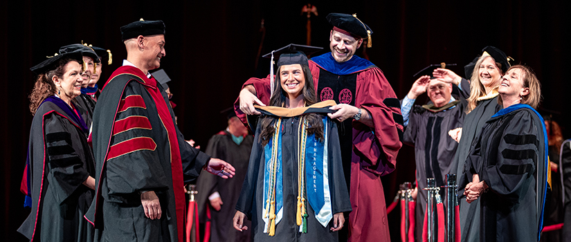 Savannah Miller smiles during HRSM's Degrees of Excellence ceremony as faculty members place her masters-style hood over her shoulders on stage.