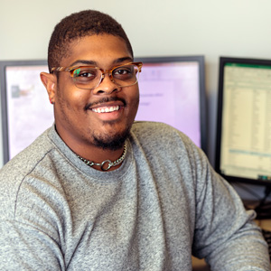 Man wearing glasses and a grey sweater sits in front of two computer screens