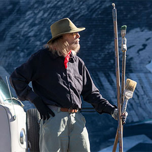 Blue Sky posed in profile with paint sticks standing in front of his mural, Tunnelvision.