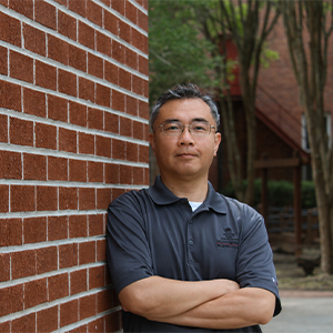 Math professor Wei Kai-Lai stands next to a brick wall on the USC Salkehatchie campus